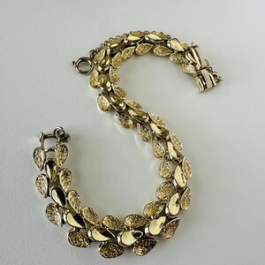 Könnte beinhalten: Ein goldfarbenes Armband mit Blattdesign. Das Armband besteht aus mehreren blattförmigen Gliedern, die miteinander verbunden sind. Die Blätter haben eine strukturierte Oberfläche und ein glänzendes Finish. Der Verschluss ist ein einfacher Haken- und Ösenverschluss.