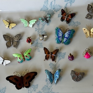 May include: A selection of butterfly brooches in diverse colours and designs. The brooches feature enamel, rhinestones, and metalwork. The butterflies are presented on a white tray with a blue floral pattern.