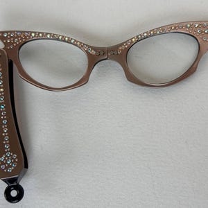 May include: Brown cat-eye glasses with clear lenses. The frames are embellished with iridescent rhinestones along the top edges. One arm is extended, also decorated with rhinestones. The glasses have a black hinge and a black circular end.