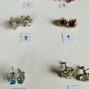 Pode incluir: Dez pares de brincos vintage de clip. Os brincos estão dispostos em filas de dois, numerados de 1 a 6. Os brincos apresentam vários estilos, incluindo designs florais, geométricos e com pedras preciosas. Os brincos são em tons prateados, dourados e pretos.