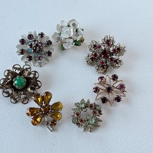 May include: A collection of vintage brooches in various floral and geometric designs. The brooches feature a range of colors, including white, gold, green, and purple, with sparkling rhinestone accents. These decorative pins are perfect for adding a touch of vintage charm.