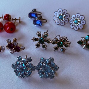 Pode incluir: Uma coleção de brincos vintage em vários estilos e cores. Inclui tachas vermelhas, brincos pendurados quadrados azuis, tachas de flores brancas e turquesas, brincos de strass roxos e transparentes, tachas de floco de neve verdes e tachas de aglomerado de flores azuis.
