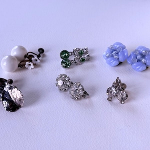 May include: A selection of vintage earrings in different designs. Features white pearl earrings with floral details, green rhinestone earrings, pale blue cluster earrings, silver leaf earrings, and rhinestone stud earrings.