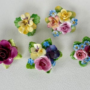 Może przedstawiać: Kolekcja pięciu ceramicznych broszek kwiatowych. Każda broszka przedstawia inny układ kolorowych kwiatów, w tym róże i bratki, z zielonymi liśćmi. Broszki są w odcieniach żółci, różu, fioletu i niebieskiego.