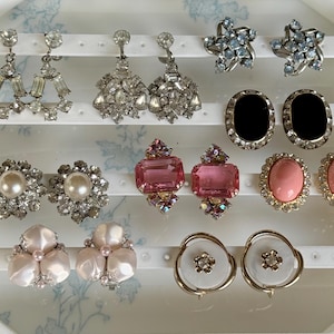 May include: A collection of ten pairs of assorted earrings displayed on a white stand. The set includes clear crystal drop earrings, star-shaped blue crystal studs, black oval cabochons, pink rectangular stones, pearl-centered floral studs, and white circular discs.