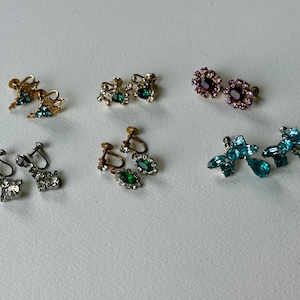 May include: An assortment of vintage clip-on earrings in various styles and colors. Includes gold-tone, silver-tone, and brass earrings with clear, green, purple, and blue rhinestones. Some earrings feature floral and bow designs.