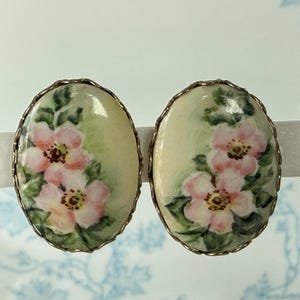 May include: Oval-shaped earrings with a hand-painted floral design. Each earring features a cluster of pink flowers with yellow centers and green leaves against a pale yellow background. The earrings are framed with a metallic border.