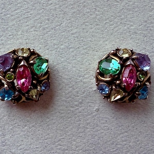 May include: A pair of vintage stud earrings with a gold-tone setting. Each earring features an arrangement of colourful gemstones, including pink, green, blue, purple, and yellow, in various shapes and sizes. The earrings have a classic, ornate design.
