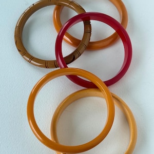 May include: A collection of six vintage bangles in various colors. The bangles are in shades of brown, orange, and red. One brown bangle has a carved design. The bangles are made of a smooth, glossy material and are arranged in a circular pattern.