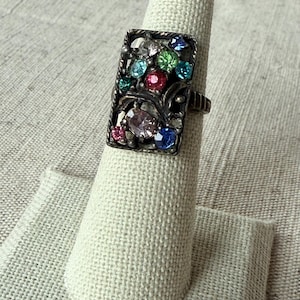 May include: A rectangular silver-toned ring with a filigree design. The ring is adorned with an array of colorful gemstones, including pink, blue, green, and clear stones. The ring is displayed on a white finger-shaped jewelry holder.