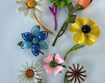 May include: A collection of colorful flower brooches in various designs. The brooches feature a daisy, a tulip, and other floral shapes in shades of white, yellow, blue, pink, and green. Each brooch has a pin on the back for easy attachment.