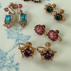 May include: A collection of vintage earrings in various styles. Includes gold-toned clip-on and post earrings with colorful gemstones. Features blue, green, pink, and purple stones in floral, geometric, and starburst designs. Some earrings have screw-on backs.