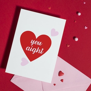 May include: A white greeting card with a red heart and the words "you aight" is displayed on a red background, next to a pink envelope. Scattered around are heart-shaped and round candies.