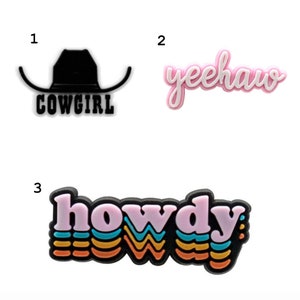 May include: Three colourful iron-on patches with western themes. The first patch is black and white and says "Cowgirl" with a cowboy hat. The second patch is pink and says "Yeehaw". The third patch is multi-coloured and says "Howdy" in a retro font.
