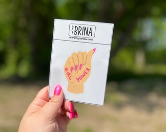 Puede incluir: Una pegatina con una mano haciendo un puño con las palabras "Girl Power" escritas en ella. La pegatina está sobre un fondo blanco con el sitio web "www.bybrina.com" impreso debajo.