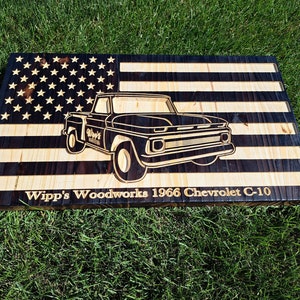 Könnte beinhalten: Eine amerikanische Flagge aus Holz mit einem schwarz-weißen Design. Die Flagge zeigt eine Silhouette eines Chevrolet C-10 Pickups aus dem Jahr 1966. Der Text "Wipp's Woodworks 1966 Chevrolet C-10" befindet sich unter dem LKW.
