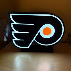 May include: A black and white illuminated sign with the Philadelphia Flyers logo. The logo features a black and white stylized wing with an orange circle in the center.
