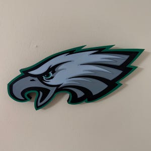May include: A wooden wall art piece featuring the Philadelphia Eagles logo. The eagle head is depicted in shades of gray, outlined in black, and accented with a green border. The artwork is mounted on a neutral-colored wall.
