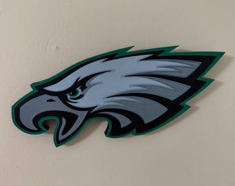 Pode incluir: Uma peça de arte de parede em madeira com o logotipo dos Philadelphia Eagles. A cabeça da águia é retratada em tons de cinza, contornada em preto e acentuada com uma borda verde. A obra de arte é montada em uma parede de cor neutra.