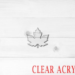 May include: A clear acrylic maple leaf cutout. The leaf is on a white wood background. CLEAR ACRYLIC is written in red letters at the bottom of the image.