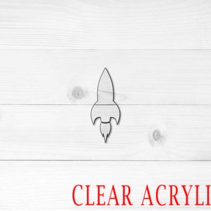 May include: A clear acrylic rocket ship cutout. The rocket ship is on a white wood background. The words "CLEAR ACRYLIC" are printed in red at the bottom of the image.