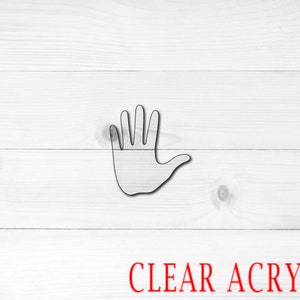 May include: A clear acrylic hand cutout with five fingers. The hand is raised with the palm facing forward. The cutout is on a white wood background.