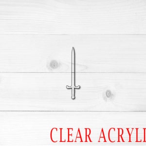 May include: A clear acrylic sword cutout. The sword has a pointed tip and a cross guard. The cutout is on a white wood background.
