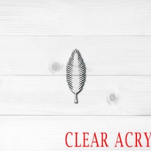 May include: A single, clear acrylic leaf cutout on a white wood background. The leaf is a simple design with a pointed tip and a stem.