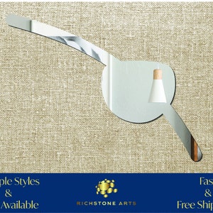 May include: A silver-colored, curved wall mirror with a rounded edge. The mirror is shaped like a crescent moon with a small, round, white light fixture hanging from the top. The mirror is on a beige, textured background.