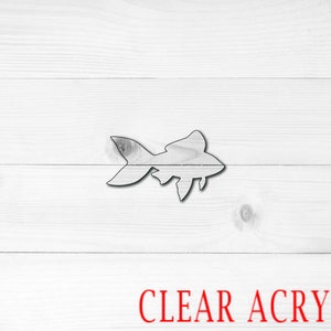 May include: A clear acrylic cutout of a fish, resembling a goldfish, on a white wood background. The text "CLEAR ACRYLIC" is printed in red below the fish.