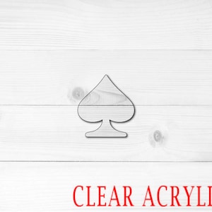 May include: A clear acrylic cutout of a spade playing card symbol on a white wood background. The text "CLEAR ACRYLIC" is printed in red below the symbol.