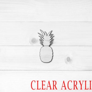 May include: A clear acrylic pineapple cutout, perfect for crafting or home decor. The pineapple is outlined in black and is shown against a white wood background. The text "CLEAR ACRYLIC" is written in red at the bottom of the image.