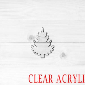 May include: A clear acrylic Christmas tree ornament in the shape of a stylized pine tree. The ornament is on a white wood background. CLEAR ACRYLIC is written in red letters below the ornament.