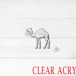 May include: A clear acrylic camel cutout, set against a white wooden background. The camel is a silhouette with visible details. The text "CLEAR ACRYLIC" is in red at the bottom.