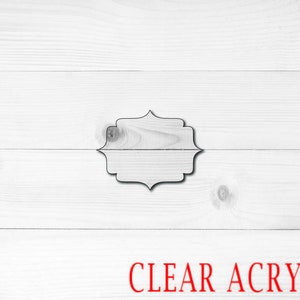 May include: A clear acrylic blank sign with a decorative frame. The sign is on a white wood background.
