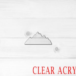 May include: A clear acrylic mountain silhouette cutout. The cutout is on a white wood background. CLEAR ACRYLIC is written in red text at the bottom of the image.