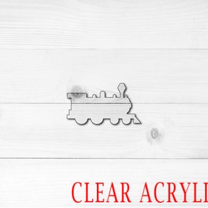 May include: A clear acrylic cutout of a train engine on a white wood background. The text "CLEAR ACRYLIC" is printed in red below the train.