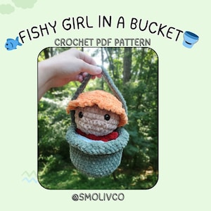 Peut inclure: Un jouet en peluche crocheté représentant une fille aux cheveux orange assise dans un seau crocheté vert. Le seau a une poignée et le jouet porte une chemise rouge. L'image est légendée "Fishy Girl in a Bucket Crochet PDF Pattern" et "@smolivco".