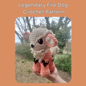 May include: A crocheted plush toy of a fire dog, with a white and orange body, black eyes, and a white mane. The toy is being held in a hand against a blurred background of trees. The text "Legendary Fire Dog Crochet Pattern" and "@heyitsnoomie" are visible below the image.