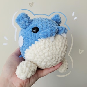 May include: A handmade plush toy in the shape of a blue and white creature with a round body, small ears, and two small white teeth. The toy has a black eye and a white spot on its side. The plush is held in a hand.