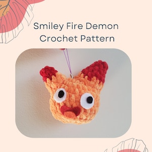 May include: A crocheted fire demon with a smiley face. The demon is orange with red accents and has large white eyes. The demon is hanging from a purple string.