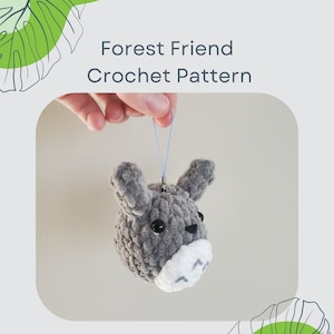 May include: A gray crocheted stuffed animal with a white face and black eyes. The animal is hanging from a blue string. The image is titled "Forest Friend Crochet Pattern".