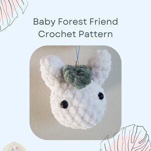 May include: White crocheted amigurumi toy with green felt ears and black eyes. The toy is attached to a blue string and is a baby forest friend crochet pattern.