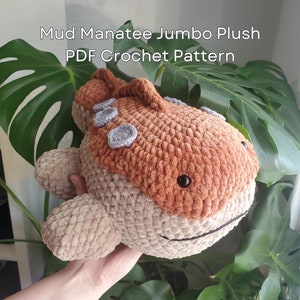 May include: Crochet pattern for a plush manatee toy. The manatee is brown and beige with a textured, fuzzy fabric. The pattern is available as a PDF download. The image includes the text "Mud Manatee Jumbo Plush PDF Crochet Pattern" and "@heyitsnoomie".