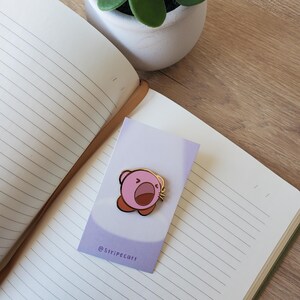 May include: A pink and gold enamel pin featuring a cartoon character with an open mouth. The pin is on a light purple card with the text "@Stripecatt". The pin is placed on a lined notebook next to a small potted plant.