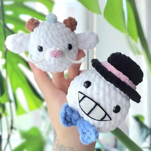 Può includere: Due peluche bianche all'uncinetto. Uno è un agnello con corna marroni e un fiore blu sulla testa. L'altro è un pupazzo di neve con un cappello nero e un papillon blu.