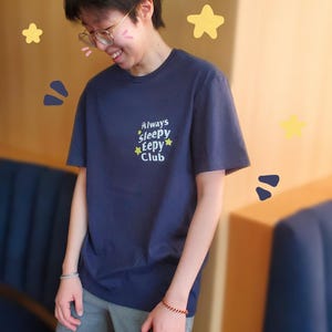 Può includere: T-shirt blu navy con la scritta "Always Sleepy Eepy Club" in un font stravagante. La maglietta è abbinata a pantaloni della tuta grigi. L'immagine presenta stelle gialle e forme blu decorative.
