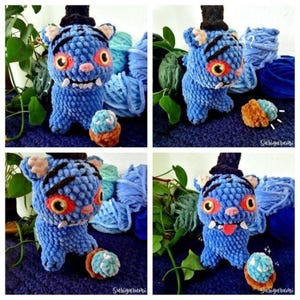 May include: A handmade blue crochet monster toy with a black top hat, orange eyes, and a pink nose. The plushie has a small tongue sticking out and is surrounded by blue yarn and a small cupcake-like toy. The toy is a unique, whimsical creation.