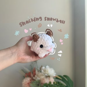 May include: A white crocheted plush toy in the shape of a small animal, featuring brown ears, black eyes, and a pink nose. The words "Snacking Snowball" are written above the toy. Decorative hearts and snowflakes are also present.