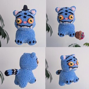 May include: A set of four images showcasing a handmade blue plush toy with a cartoonish tiger-like design. The toy has large yellow eyes, a toothy grin, and black accents. One version has a small black hat. The toy is holding a small brown and pink object in one image.